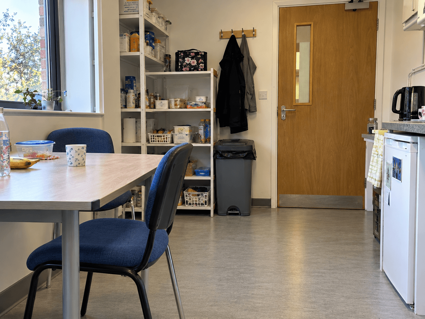 An unobstructed staff-room chair and table, with a worker's lunch bag on a shelf and a clear route to the door.