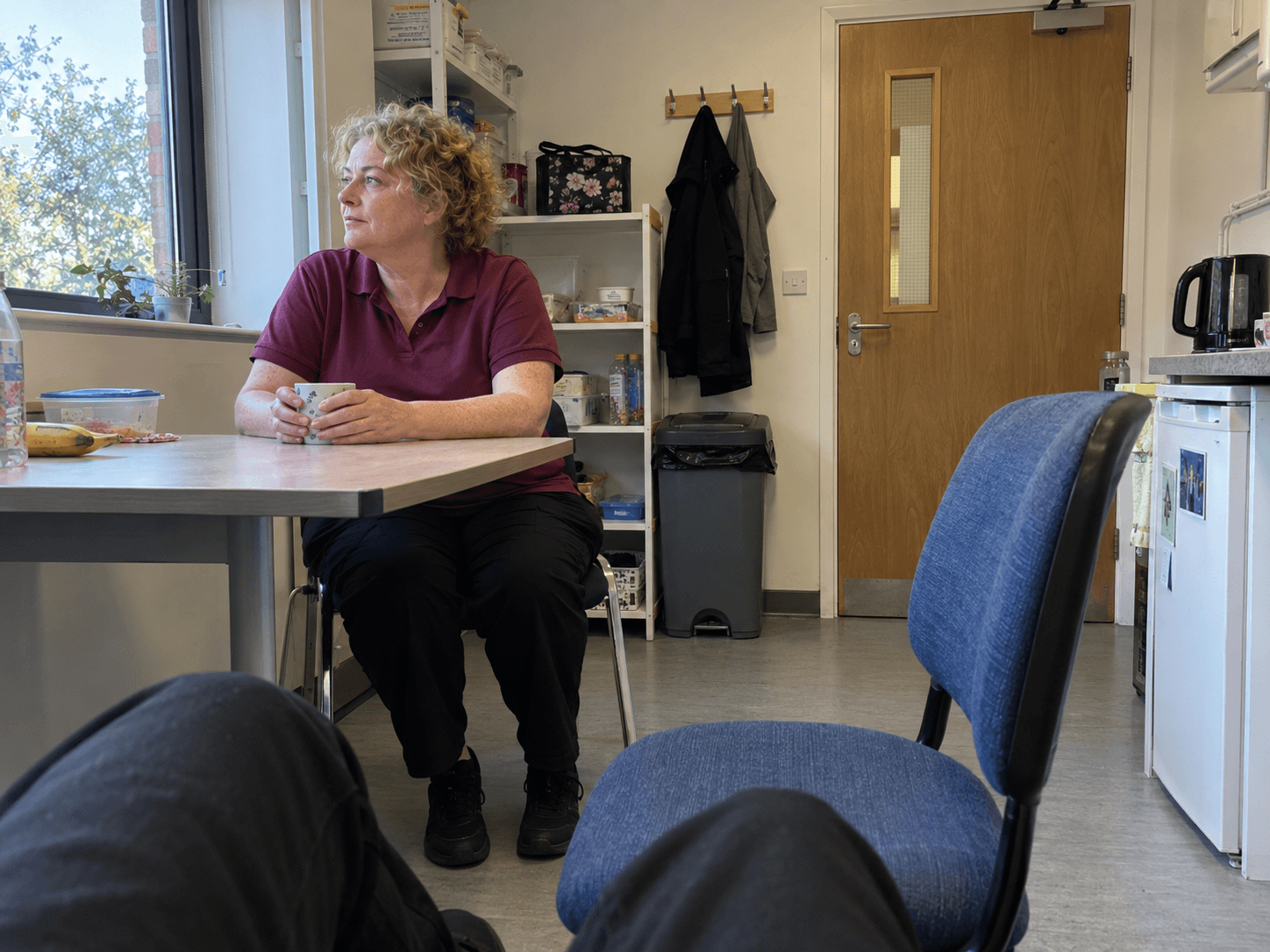 A shop worker in a plain burgundy polo sitting at a small staff-room table during a break, with both feet supported on the floor.