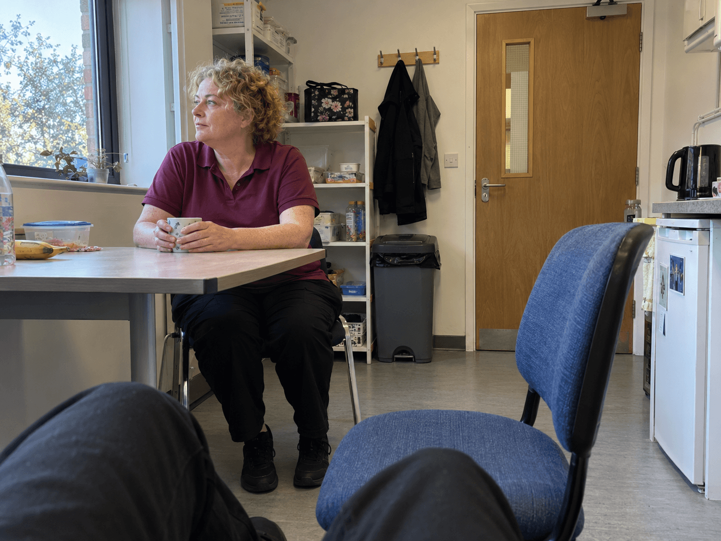 A shop worker in a plain burgundy polo sitting at a small staff-room table during a break, with both feet supported on the floor.
