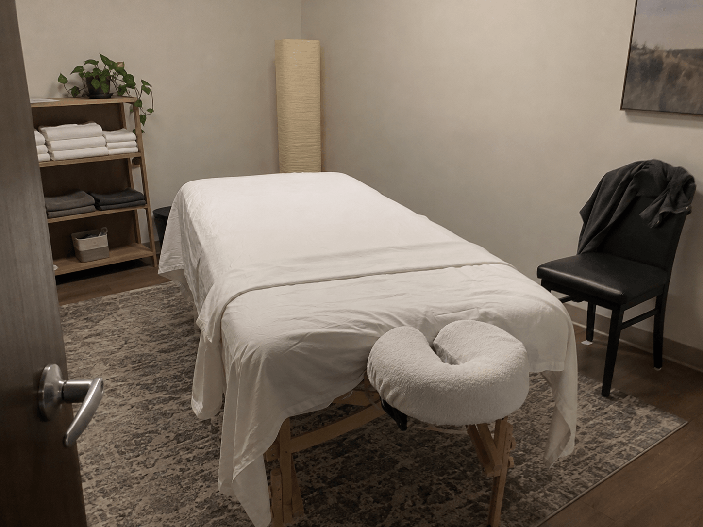 An empty treatment room with a covered massage table, folded towels and a jacket on a chair.