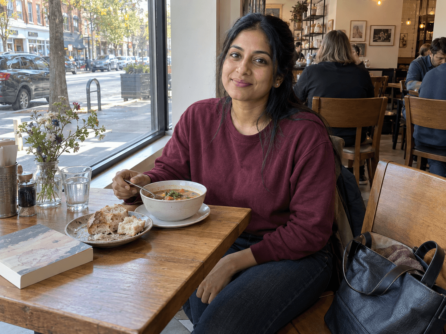 Priya seated by a cafe window with a bowl of soup and bread, looking up with a small smile.