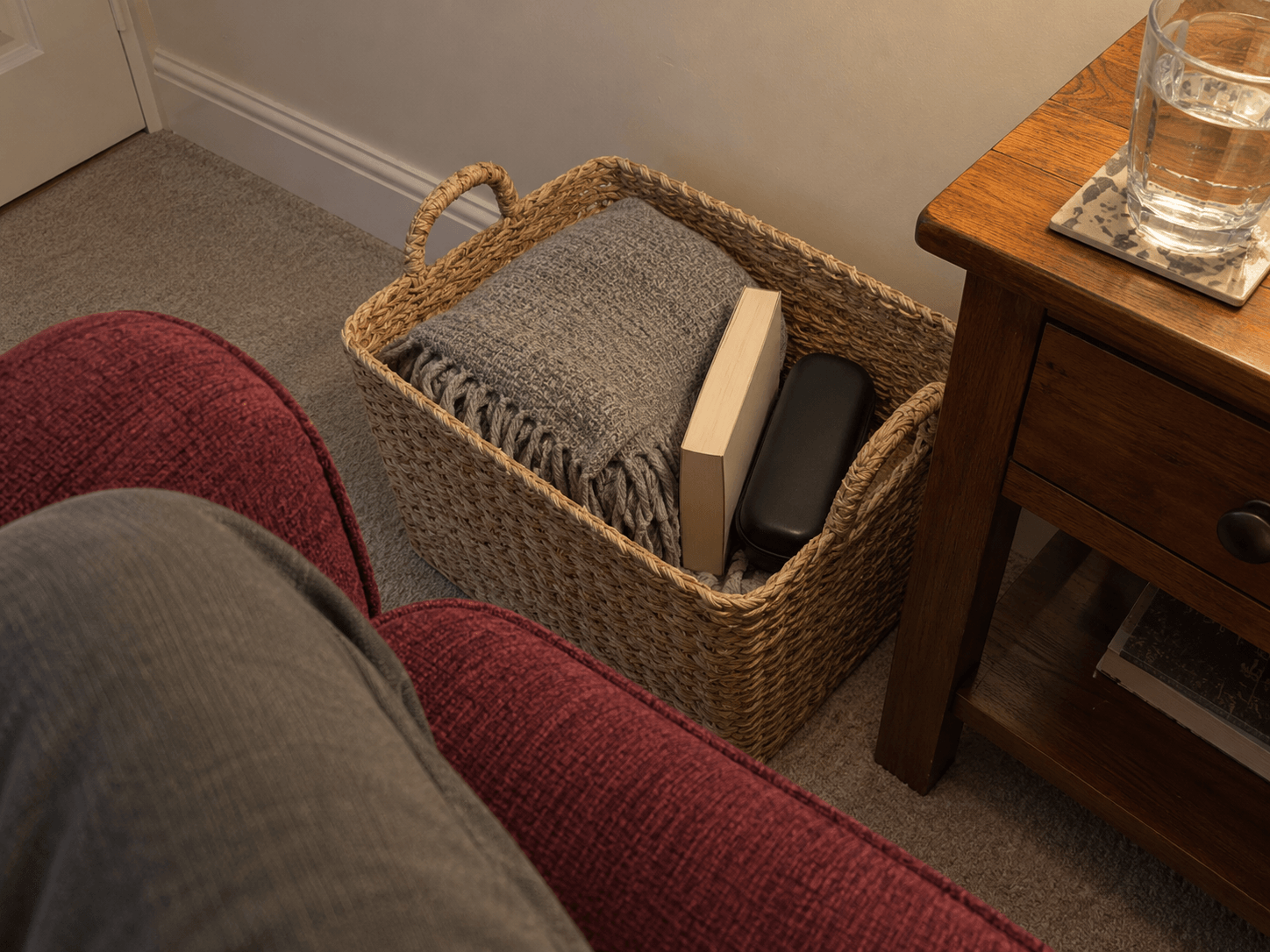 A seated view of a woven basket beside an armchair, holding a grey blanket, a paperback and a glasses case, with a glass of water on a separate table.