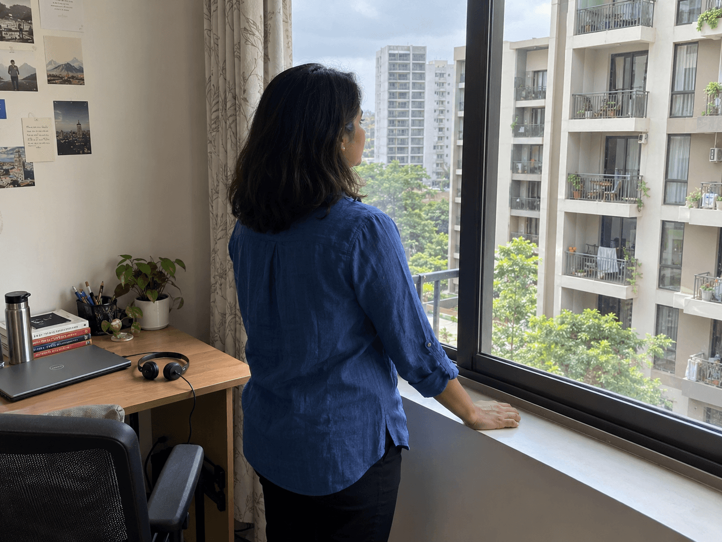 A woman in a blue shirt standing at her apartment window after a call, her headset resting beside a closed laptop on the nearby desk.