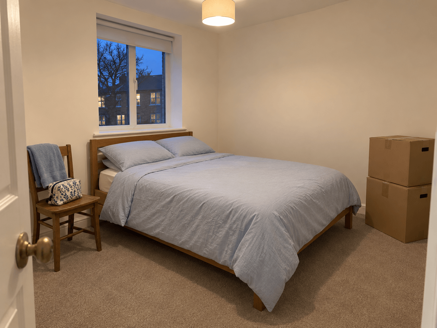 A made bed with a towel and wash bag on a nearby chair, while a few closed moving boxes remain along the opposite wall with the doorway clear.