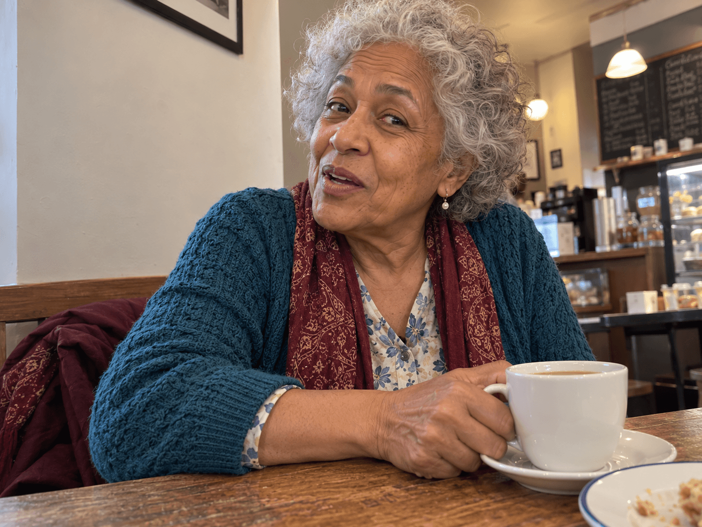 Aunt May speaking across a cafe table, photographed closely by Leila, with her tea and burgundy scarf partly in view.