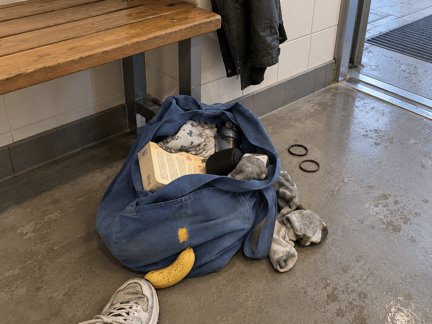 A repaired blue tote beside an entrance bench with a paperback, socks and a banana by a shoe.