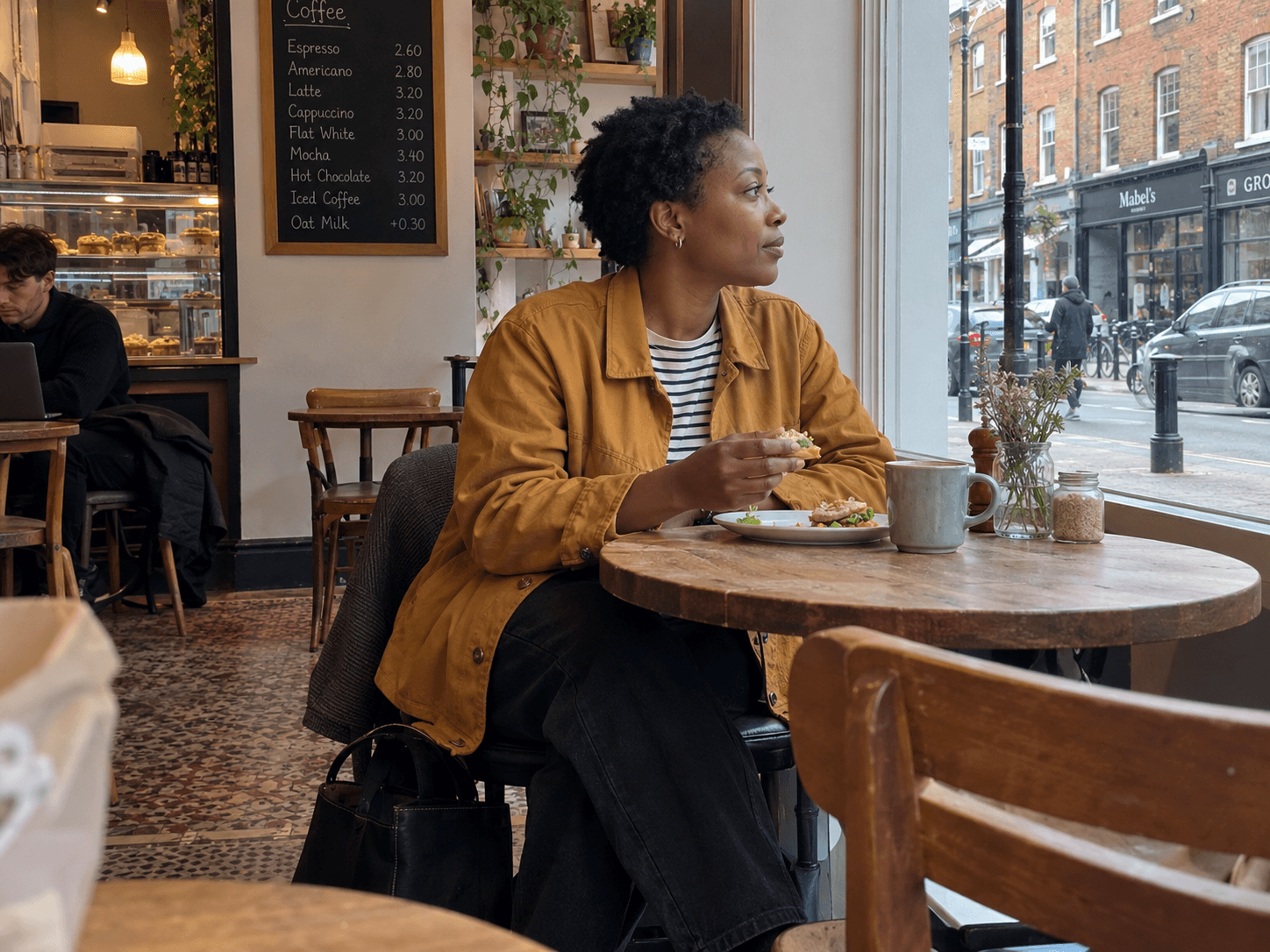 Nadine, a Black woman in her early forties with short natural hair and a mustard jacket, sitting by a neighbourhood cafe window with a sandwich and a practical work bag under the table.