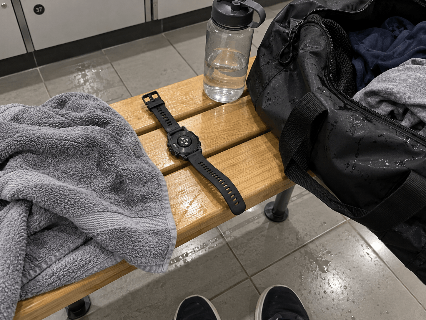 A face-down sports watch on a changing-room bench beside a grey towel, water bottle and gym bag.