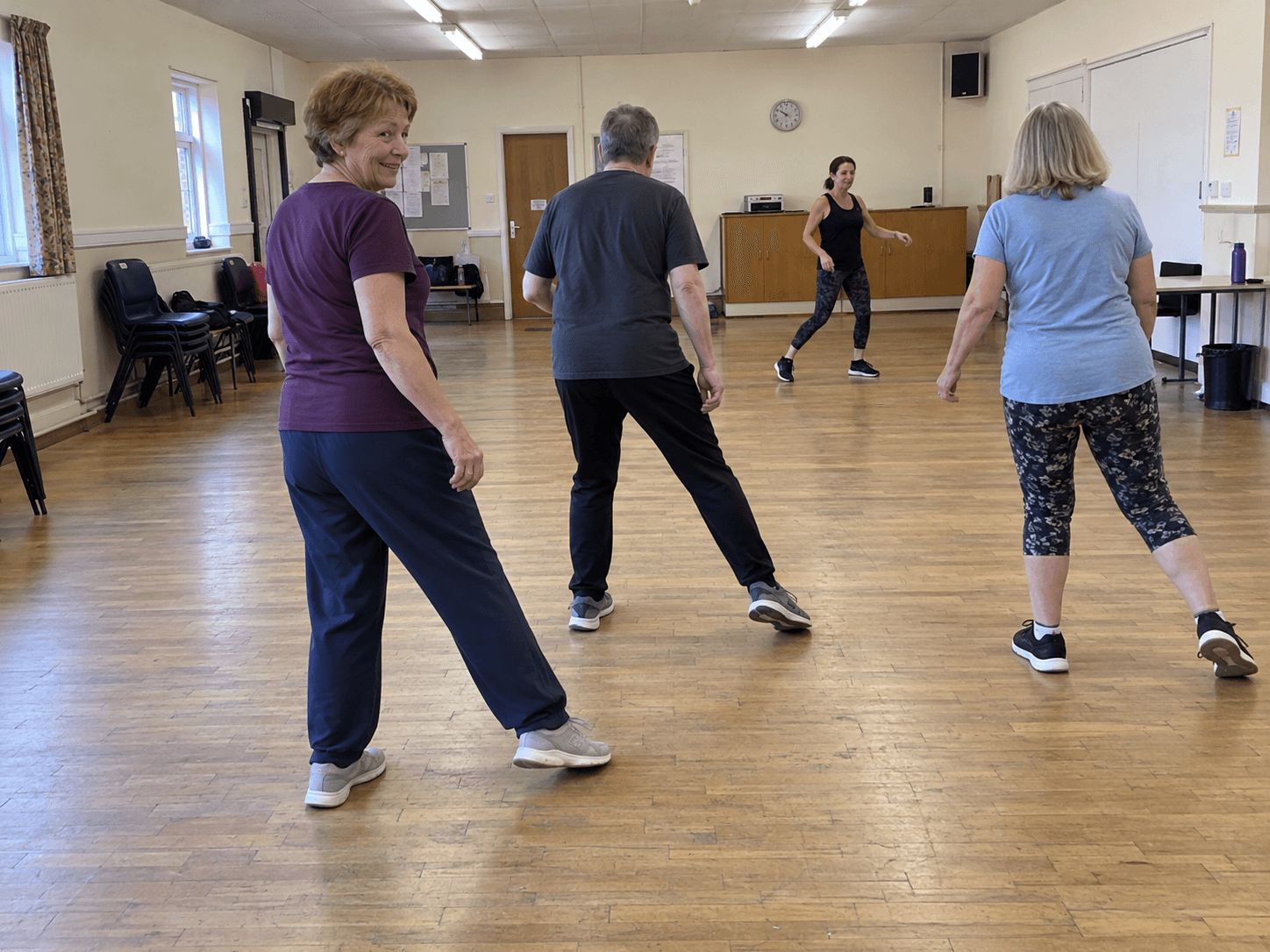 Marina in a plum T-shirt and comfortable trousers practising a simple side step in a plain community-room dance class.
