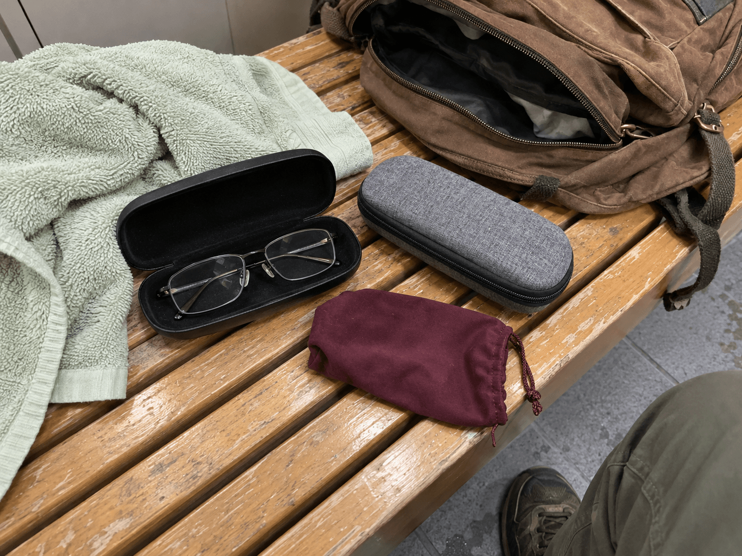 Three different glasses cases beside a green towel and an open brown backpack on a worn bench.