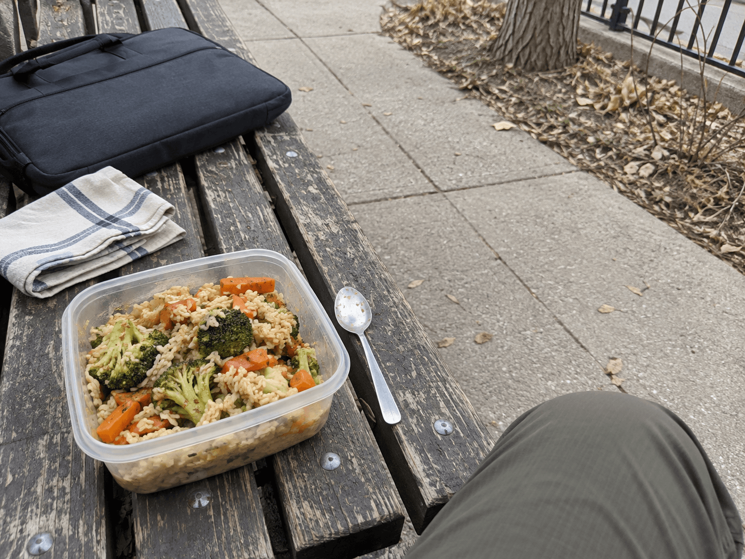 A container of rice and vegetables with a small teaspoon on a worn outdoor bench beside a work bag.
