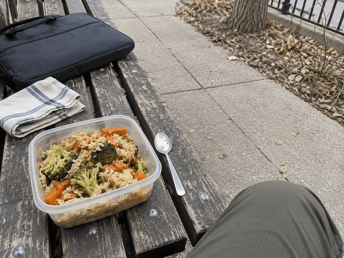 A container of rice and vegetables with a small teaspoon on a worn outdoor bench beside a work bag.
