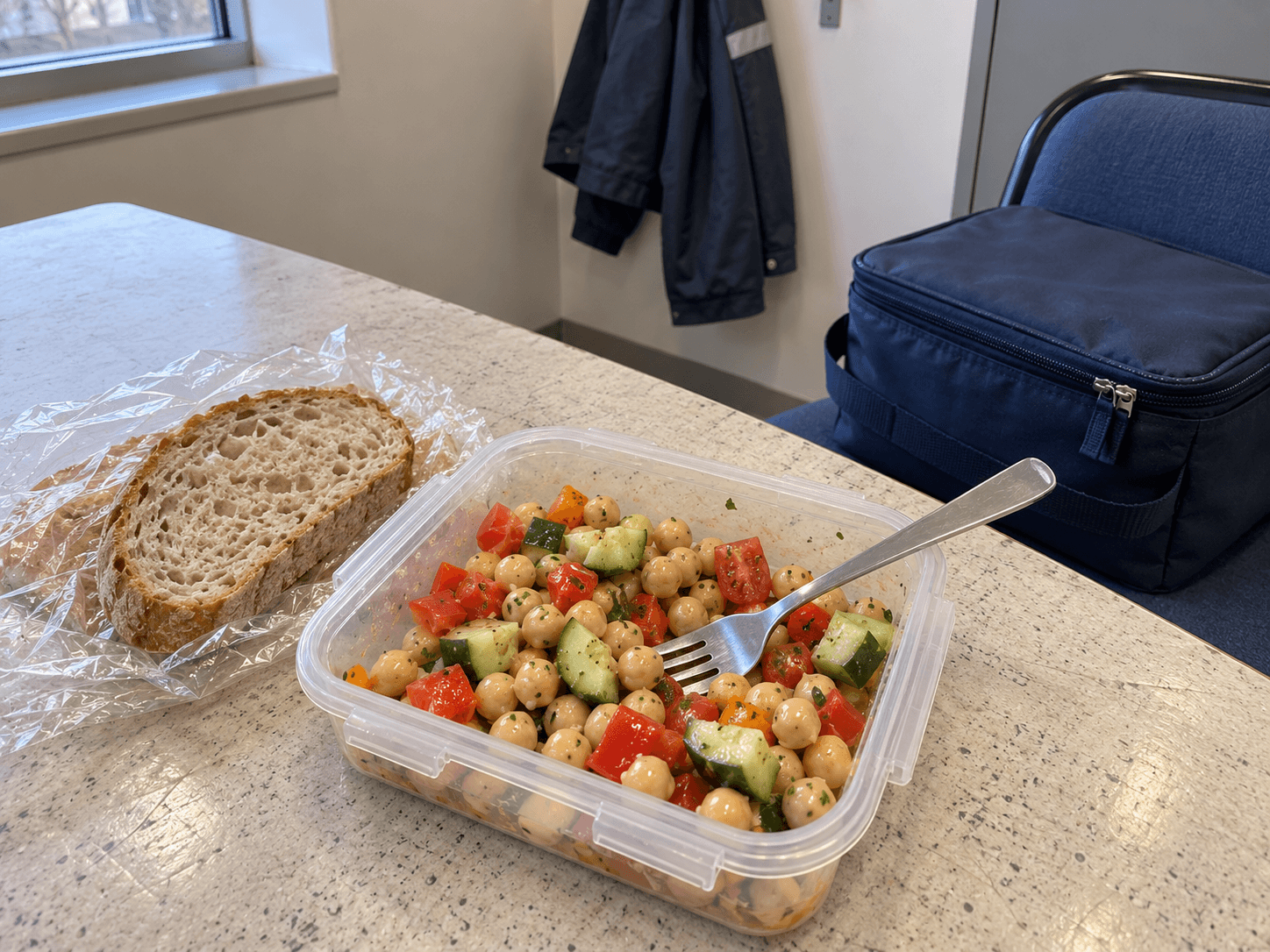 A chickpea and chopped vegetable salad, bread and a fork on a plain staff-room table, with the closed insulated lunch bag beside the chair.