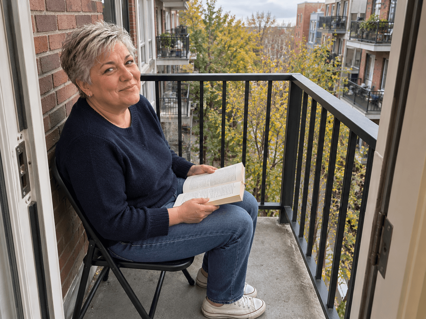 Bea seated in a simple folding chair on a small apartment balcony, reading a paperback with the door and walking space clear.