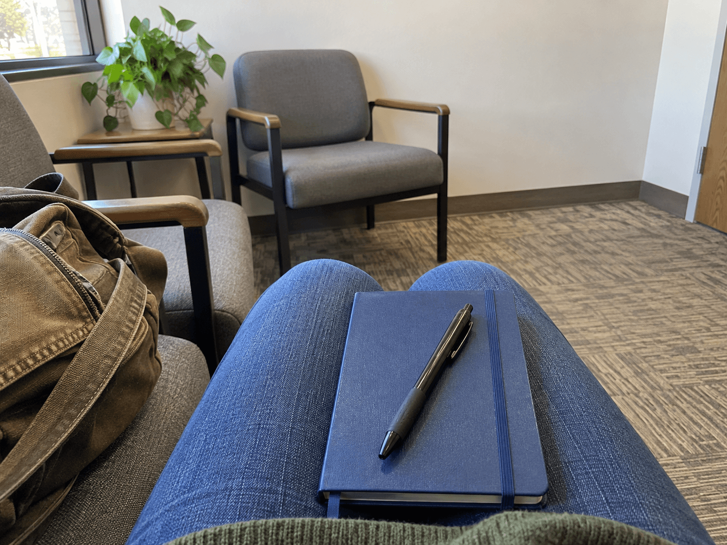 A closed blue notebook and pen on clothed knees in a simple waiting area.