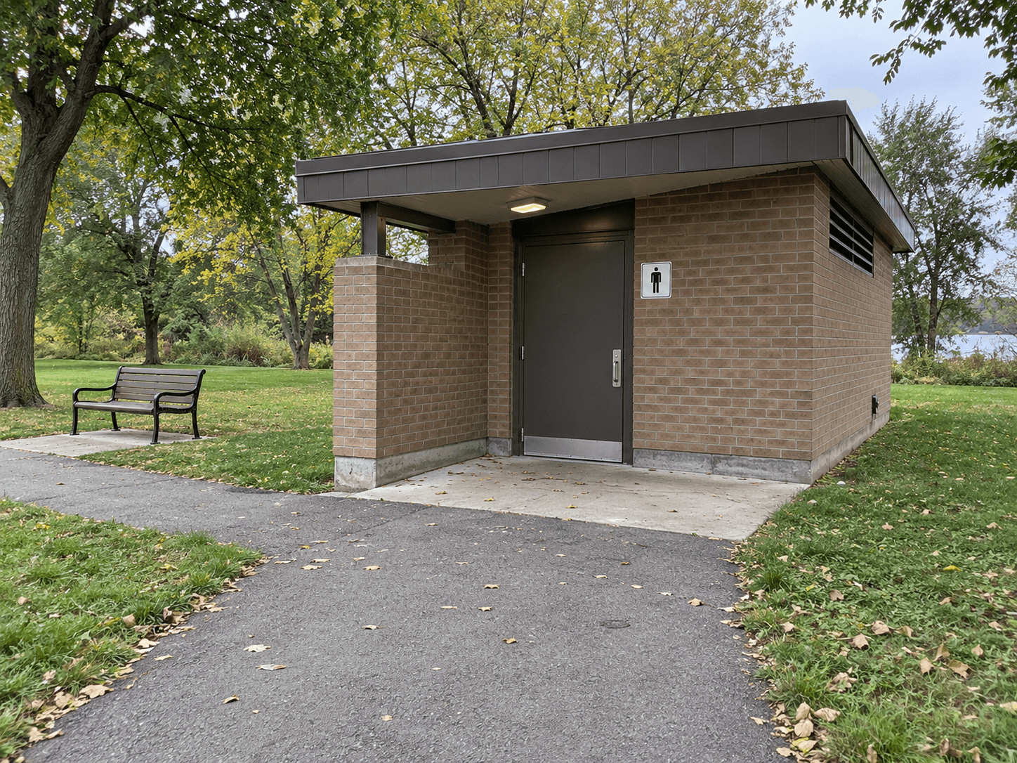 A small brick public washroom building beside a paved park path, with its entrance and a nearby bench visible.