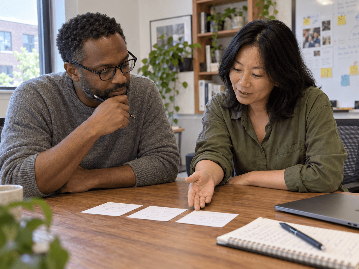Jo in an olive shirt at a small office table with a colleague, looking together at three simple task cards rather than at a laptop screen.