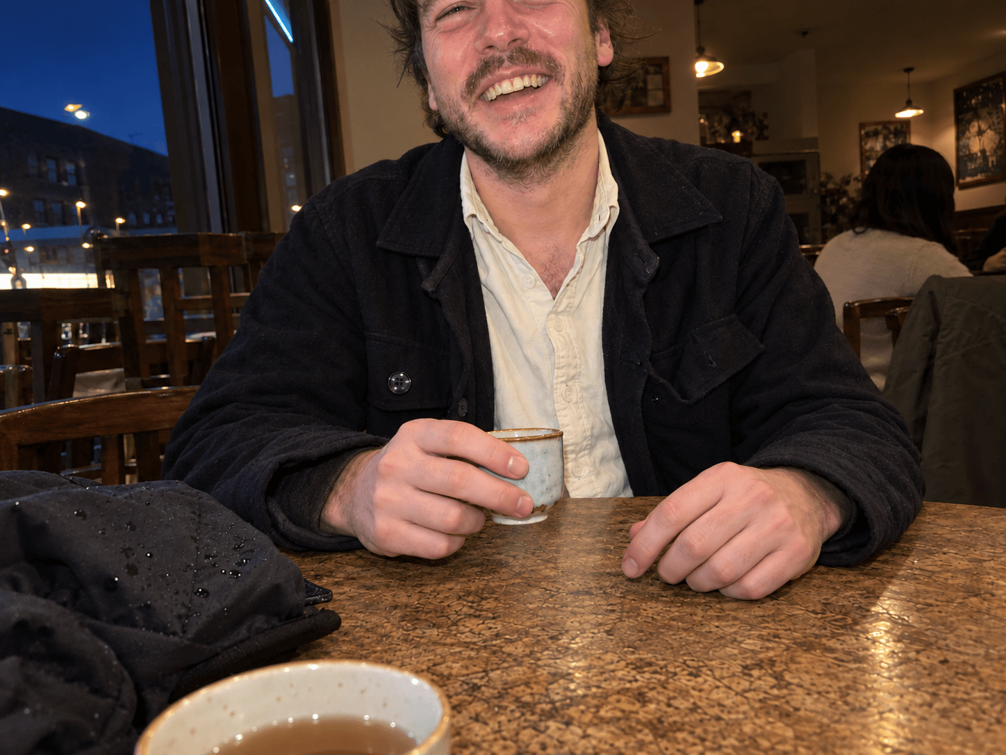 Ben laughing across a cafe table with a tiny tea cup in a casually cropped phone photograph.