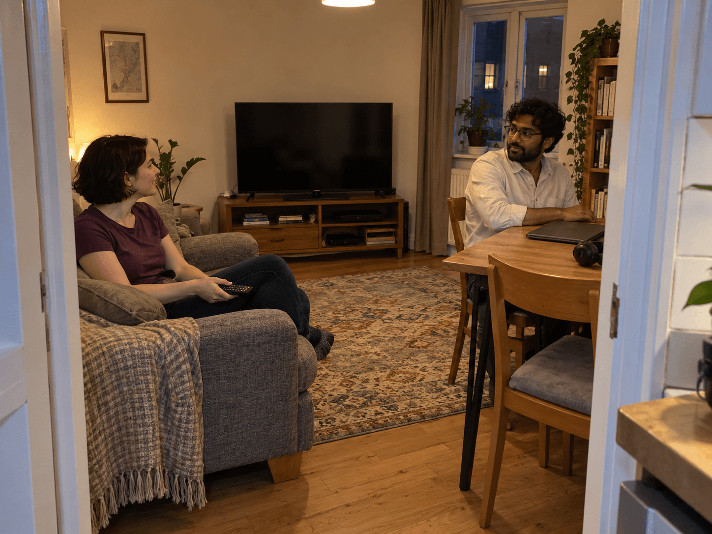 Two housemates talking across their shared sitting room, one on the sofa with a television remote and one at a table beside a closed laptop and headphones.