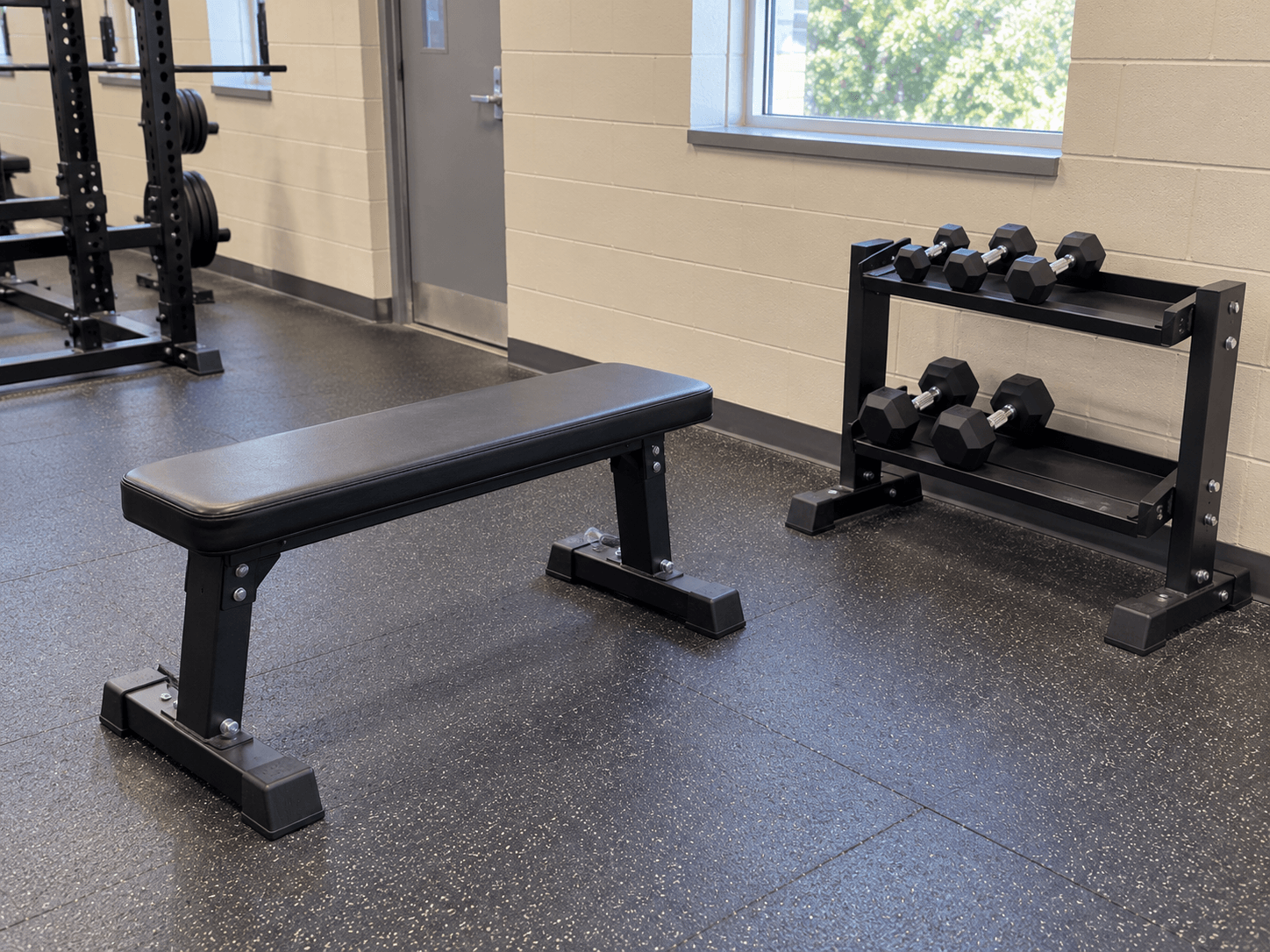 An empty exercise station with a stable bench, clear floor space and a pair of light dumbbells stored on a low rack.