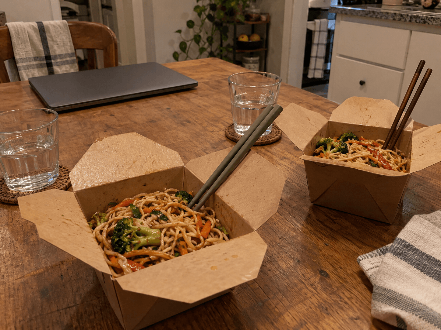 Two plain takeaway containers of noodles and vegetables on a kitchen table.