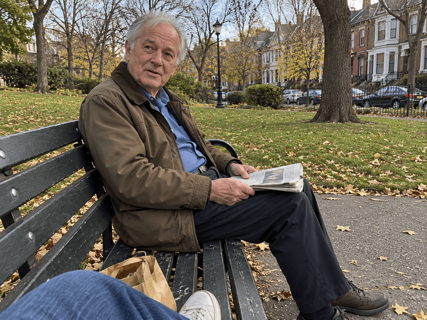 An older man in a brown jacket seated at one end of a public bench, holding a folded newspaper while looking toward his neighbour beside the camera.