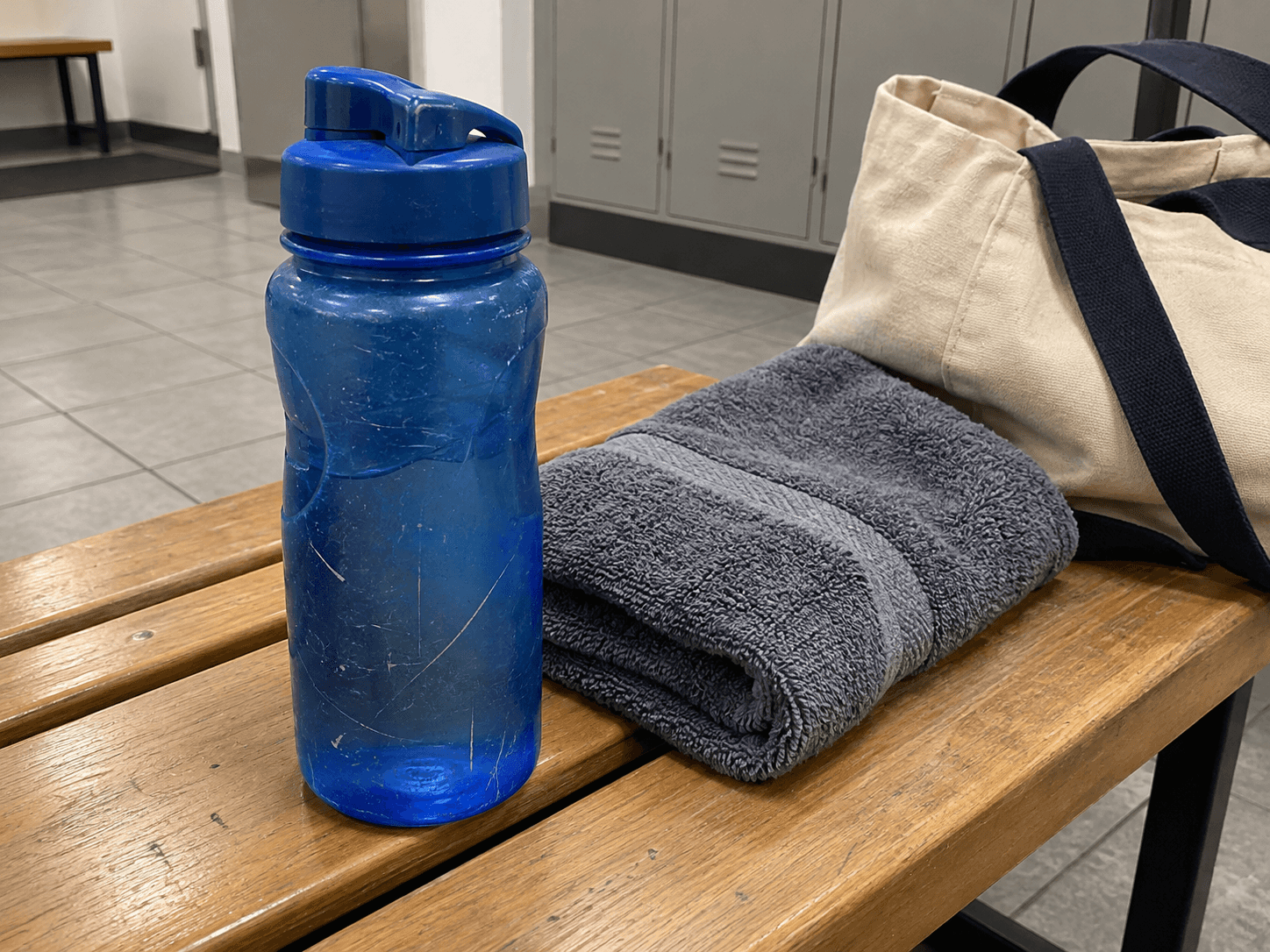A blue reusable water bottle beside a folded grey towel and canvas bag on a changing-room bench.