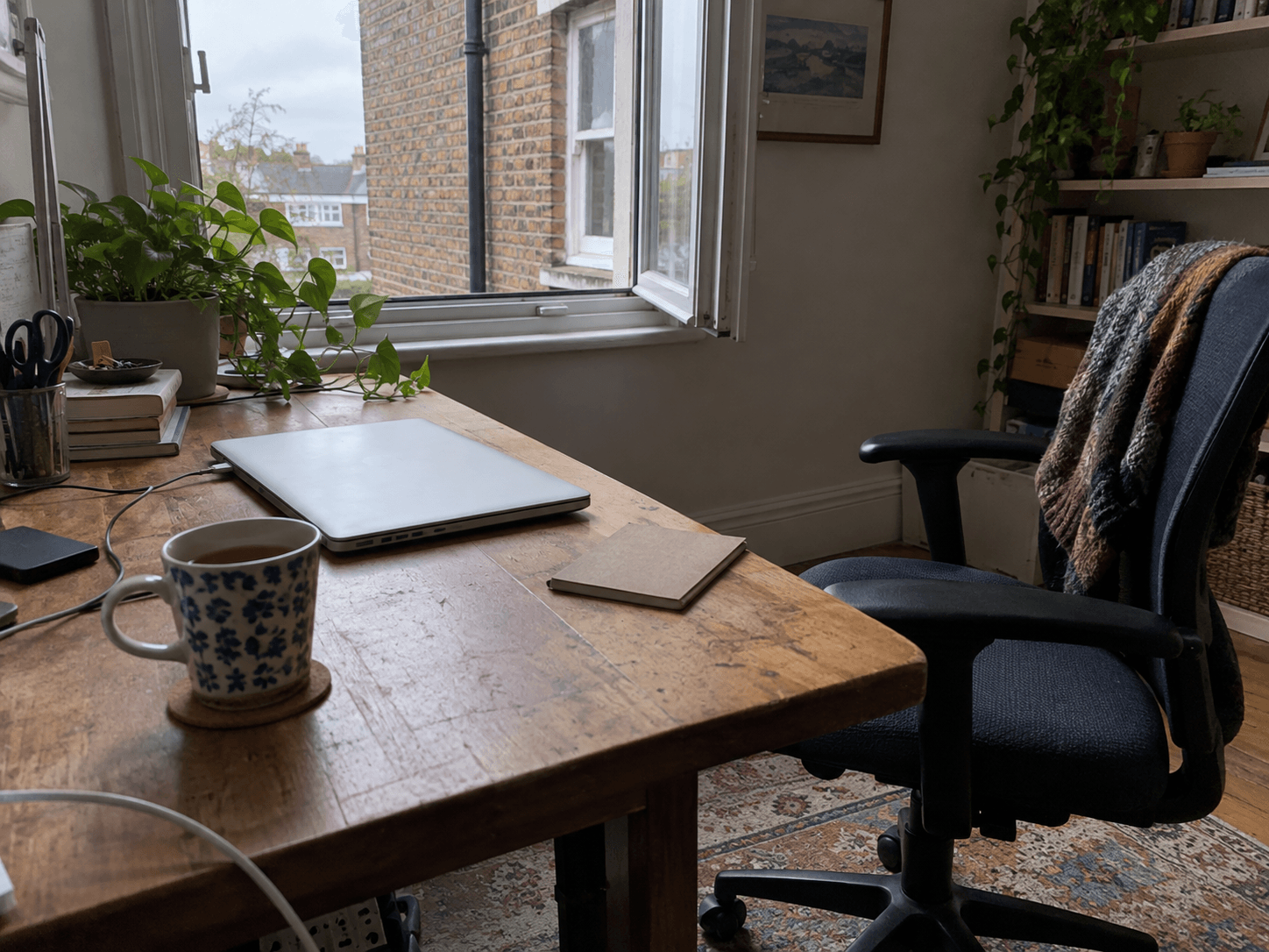 A closed laptop and mug on an ordinary desk beside an open window.
