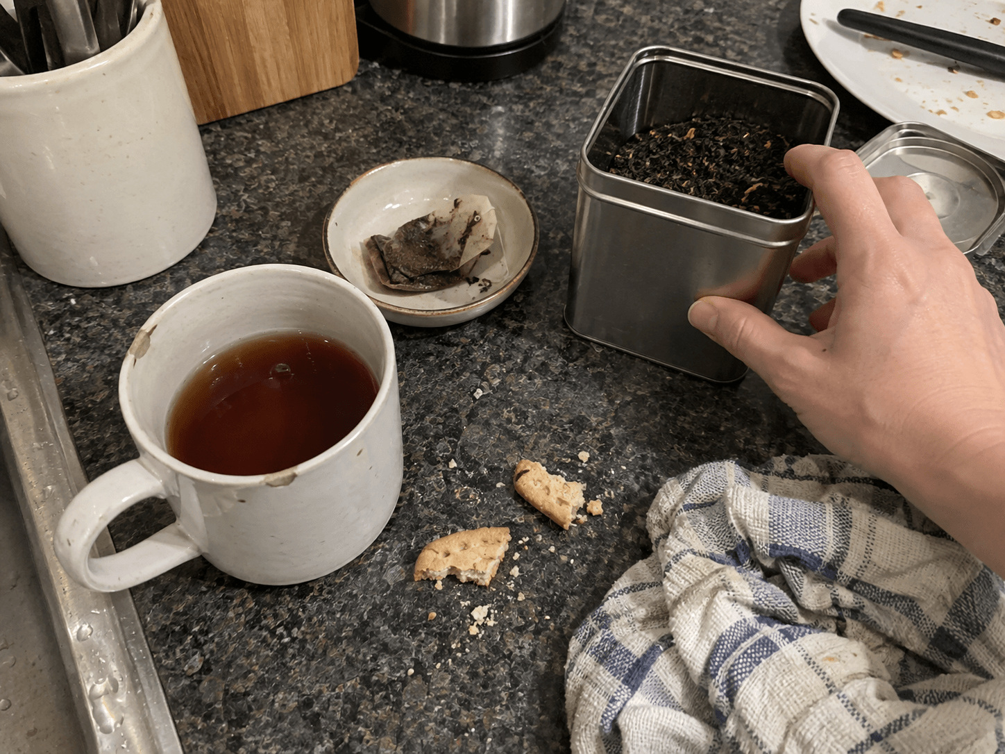 A mug of dark tea, a used teabag and an open tin on an ordinary kitchen counter.