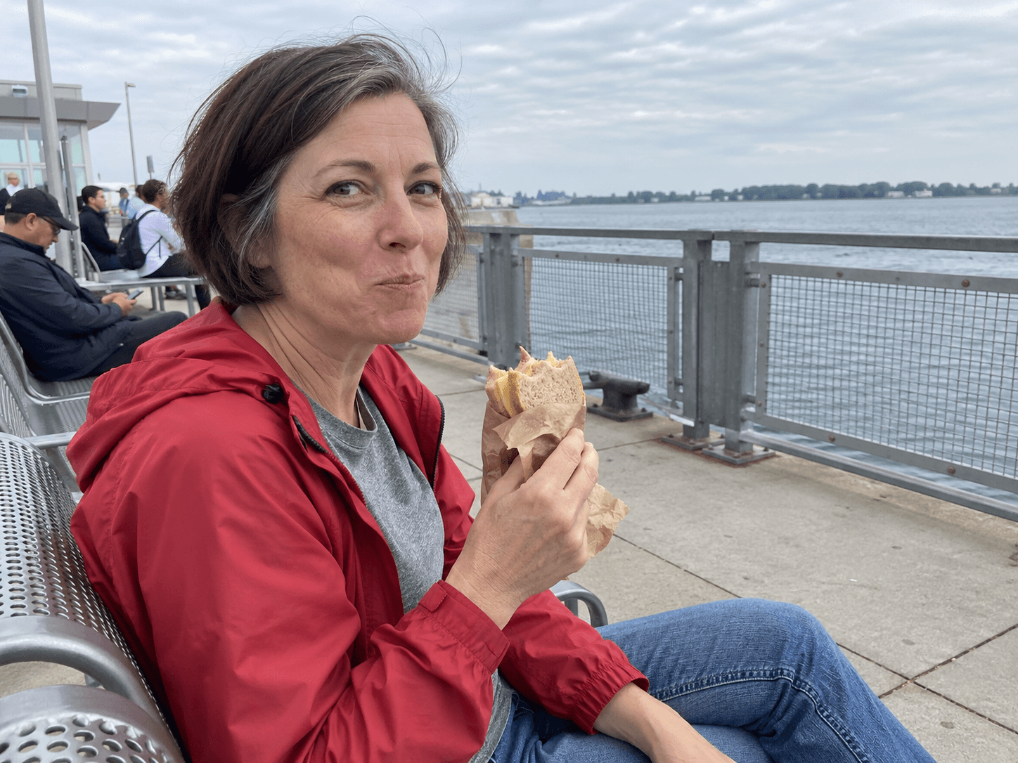 A woman in a red windbreaker eating a wrapped sandwich in a ferry-terminal waiting area, with the harbour beyond the rail.
