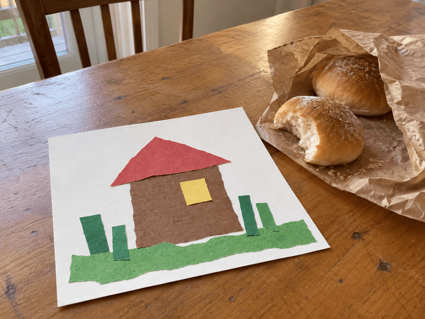 A small uneven paper collage of a house and garden beside a partly eaten bun on its paper bag.