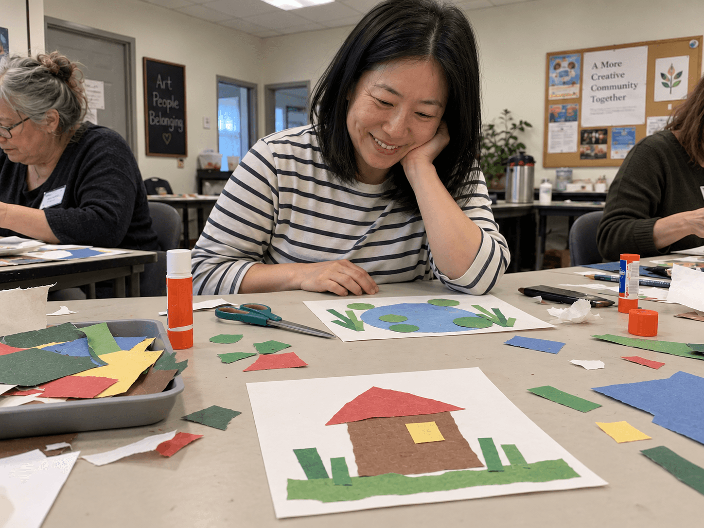 Rina smiling at a small simple paper house on a community art table, with her own paper pond beside it.
