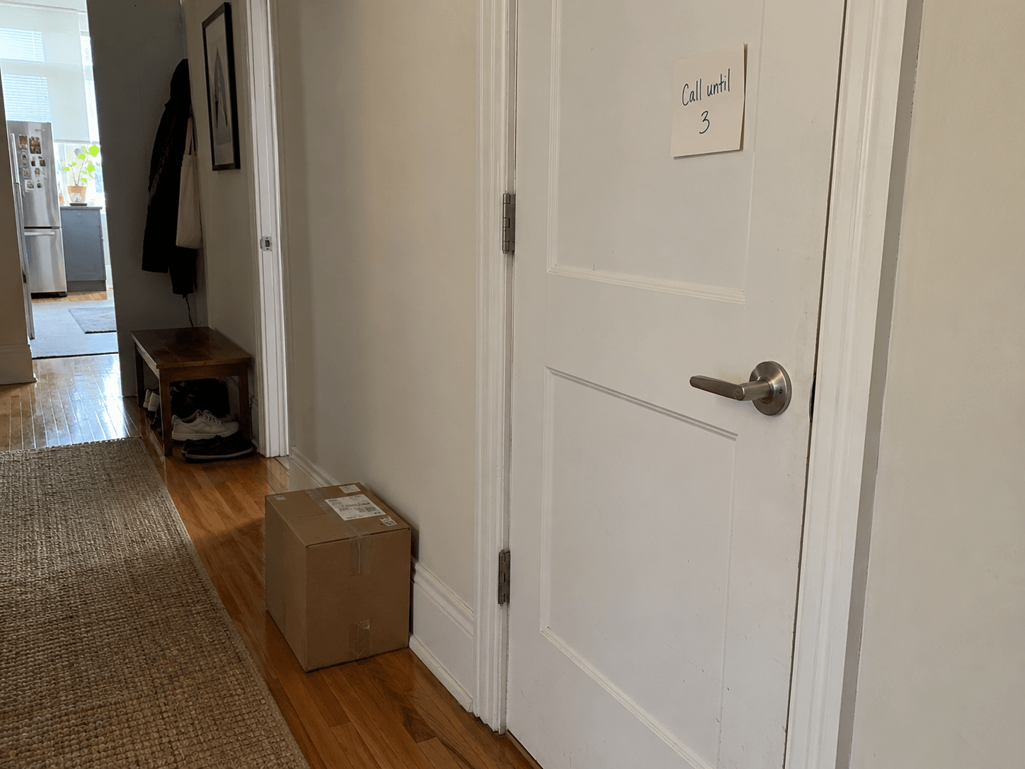 A closed ordinary bedroom door in a shared apartment with a small handwritten card saying Call until 3, and a parcel placed safely beside the wall.