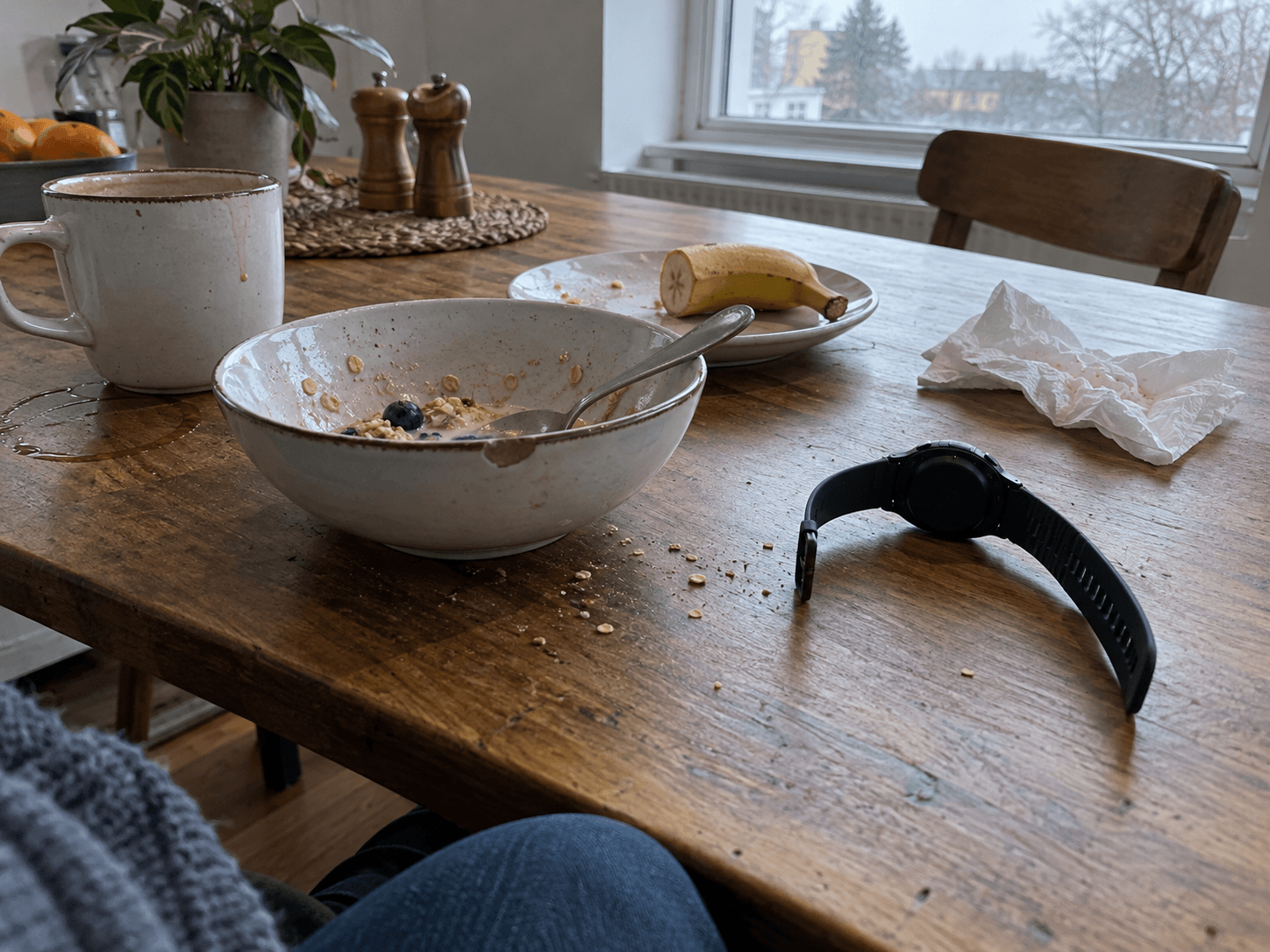 A fitness watch face down beside a cereal bowl, banana and mug on a breakfast table.