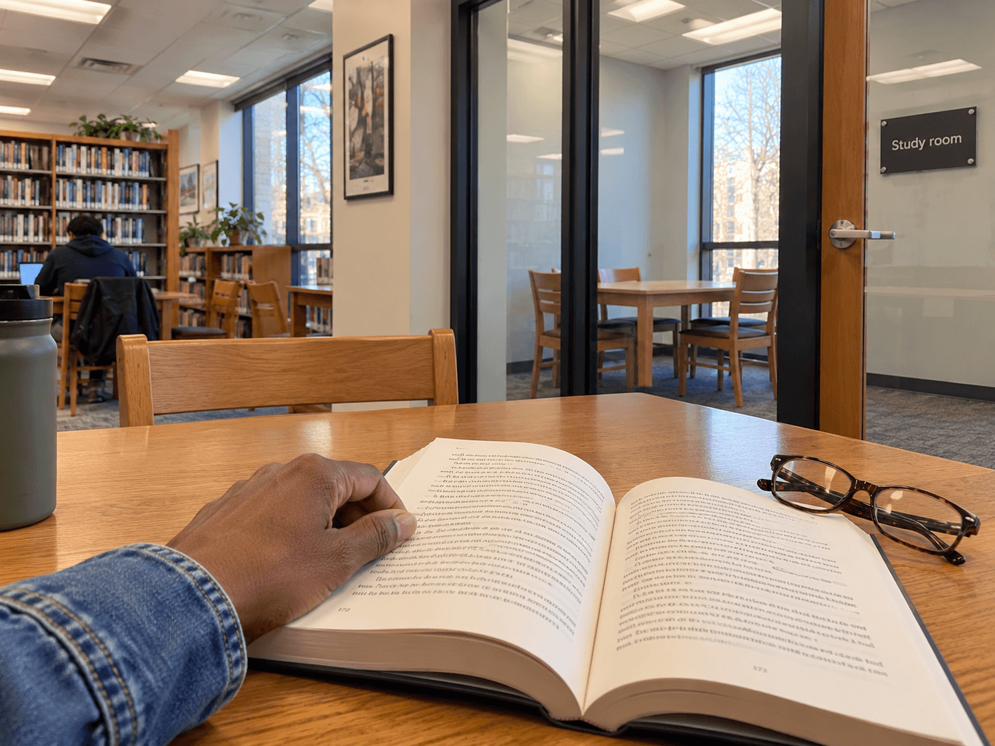 A book and reading glasses on a library table, with a separate glass-fronted study room beyond the aisle.