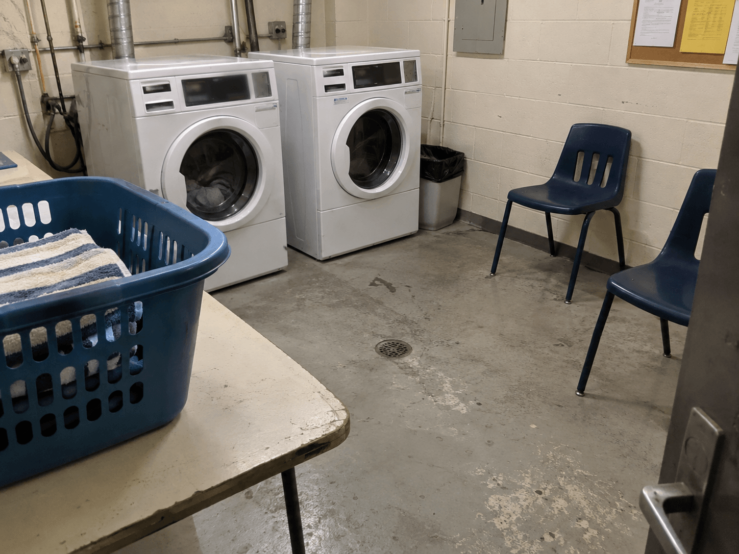 A blue laundry basket and two plastic chairs in an apartment laundry room.