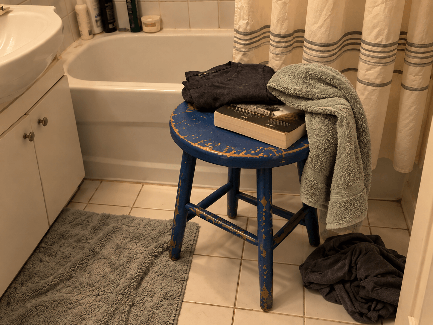 A chipped blue stool holding a paperback, folded clothes and a pale green towel beside the bathtub, with a dark T-shirt on the floor.